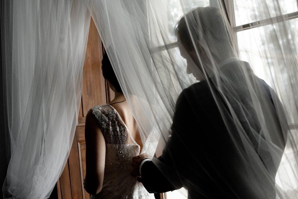 A groom helps his bride prepare, captured through a delicate veil in an intimate indoor setting.